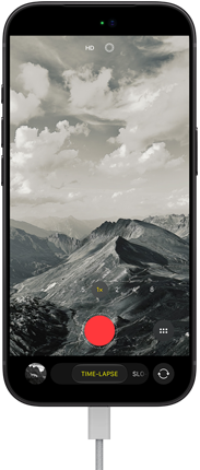 „iPhone 18 Pro“, priekinė pusė iš išorės, ekrane kalnuotas kraštovaizdis su kameros režimo juosta, pasirinktas „Time-Lapse“ režimas, prie įkrovimo prievado prijungtas maitinimo laidas