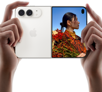 „iPhone Duo“, žvaigždžių balta spalva, laikomas dviem rankomis horizontaliai, išlankstytas; kairėje pusėje matomos 48 MP „Dual Fusion“ galinės kameros, dešinėje išoriniame ekrane rodomas fotografuojamas žmogus, kuris fotografavimo metu mato savo veidą naudodamas „Duo Preview“