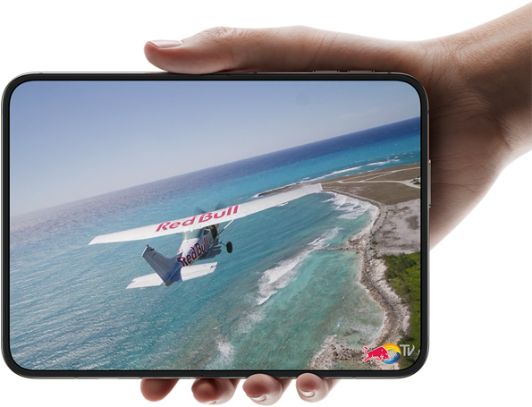 „iPhone Duo“, laikomas viena ranka, atidarytas horizontaliai, visiškai išlankstytas, vidiniame ekrane rodoma nuotrauka su virš paplūdimio skrendančiu lėktuvu.