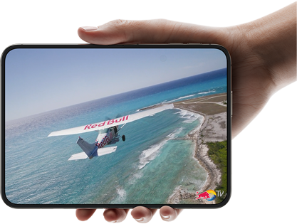 „iPhone Duo“, laikomas viena ranka, atidarytas horizontaliai, visiškai išlankstytas, vidiniame ekrane rodoma nuotrauka su virš paplūdimio skrendančiu lėktuvu.