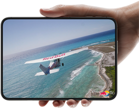„iPhone Duo“, laikomas viena ranka, atidarytas horizontaliai, visiškai išlankstytas, vidiniame ekrane rodoma nuotrauka su virš paplūdimio skrendančiu lėktuvu.